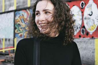 Vrouw met lang zwart los haar en zwarte trui staat voor graffiti muur en is blij met pijn in mond door stress behandeling.