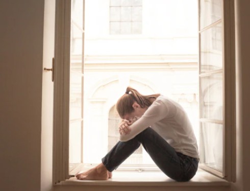 Jonge vrouw met wit shirt en donker legging zit voor groot raam en vraagt zich af wat de spierzwakte door stress symptomen zijn.