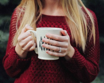 Vrouw met blond haar en rode trui heeft beker warm drinken in haar hand en vraagt zich af wat de koud door stress oorzaak is.