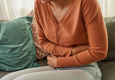 Vrouw met bruin shirt zit op bank met armen om haar buik omdat ze last heeft van rommelende darmen stress symptomen.